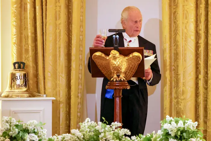En eldre herre i formell antrekk med medaljer taler ved en talerstol med et ørneemblem, holder et glass og notater, omgitt av et blomsterarrangement