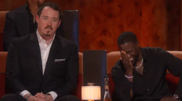 Shane Gillis og Kevin Hart sitter i et publikum; Shane er i dress og ser alvorlig ut, mens Kevin i en uformell skjorte ler og dekker ansiktet med hånden