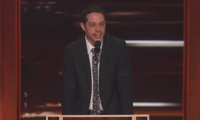 Pete Davidson i dress og slips taler ved et podium på scenen