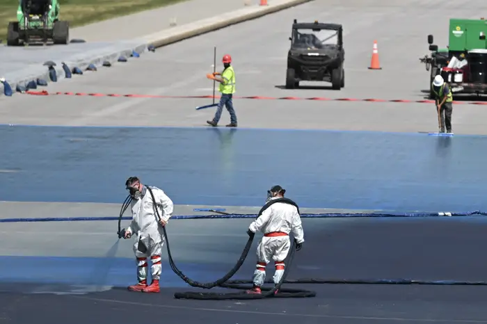 Arbeidere i beskyttelsesdrakter sprayer stoff på en stor overflate på et utendørs område. Sikkerhetsbarrierer og maskineri er synlige i bakgrunnen