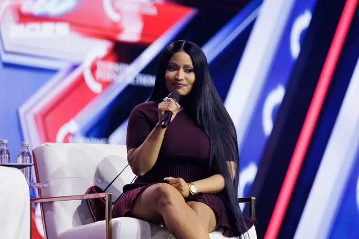 Nicki Minaj i en elegant, kortermet kjole sitter på scenen med en mikrofon og snakker på et offentlig arrangement