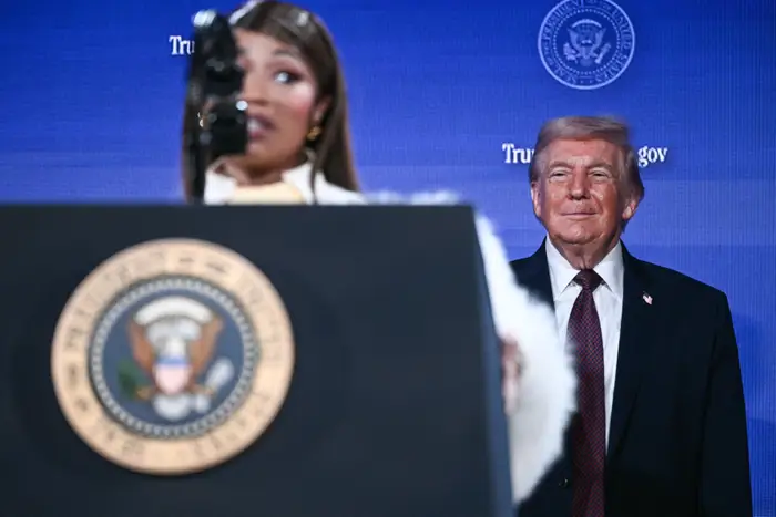 Nicki Minaj taler ved et podium med en presidentsegl, mens Donald Trump står i bakgrunnen og observerer