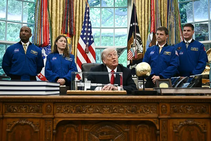 En gruppe NASA-astronauter står rundt et skrivebord i det ovale kontoret mens en mann i dress sitter og snakker