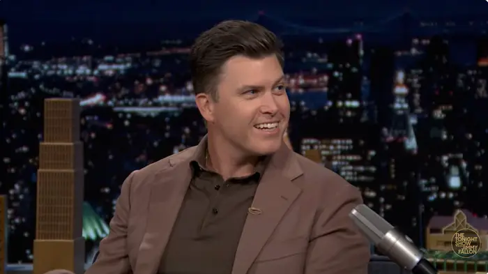 Person i dress som smiler under et TV-talkshow-intervju med et bybilde som bakteppe