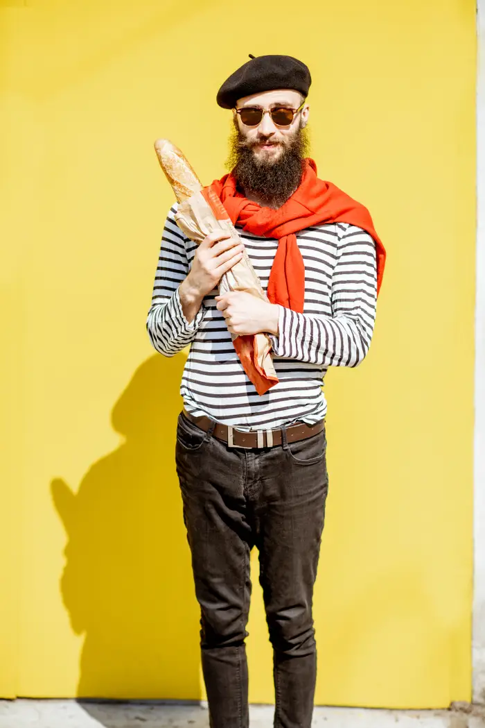 En mann med skjegg og solbriller holder en baguette. Han har på seg en beret, stripet skjorte og rødt skjerf, og står foran en vanlig bakgrunn