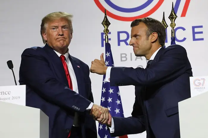 To menn i dress står på en scene under et G7-arrangement og håndhilser. En ser ut til å peke. Et G7 Frankrike-skilt er synlig i bakgrunnen