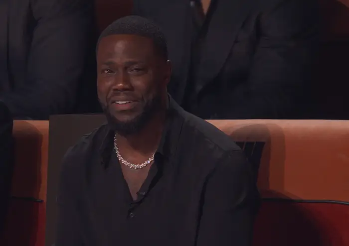 Kevin Hart i mørk skjorte, smilende, sitter i et publikum