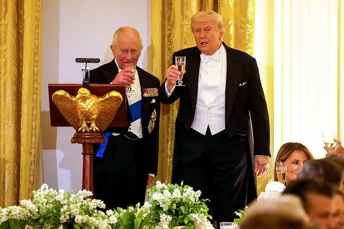To menn i formelt kveldstøy hever brillene på et podium under et arrangement. En kvinne er delvis synlig ved bordet nedenfor
