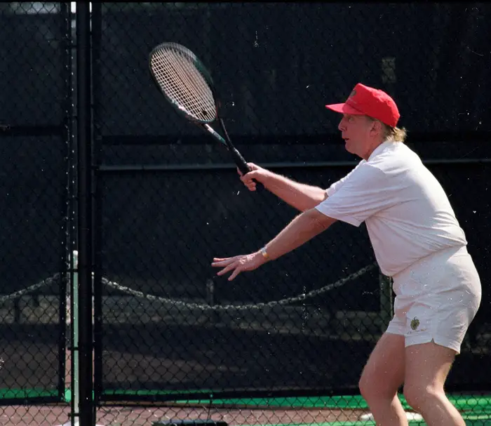 Person som spiller tennis, iført et hvitt antrekk og caps, forbereder seg på å gi et skudd på en bane med et kjettinggjerde i bakgrunnen