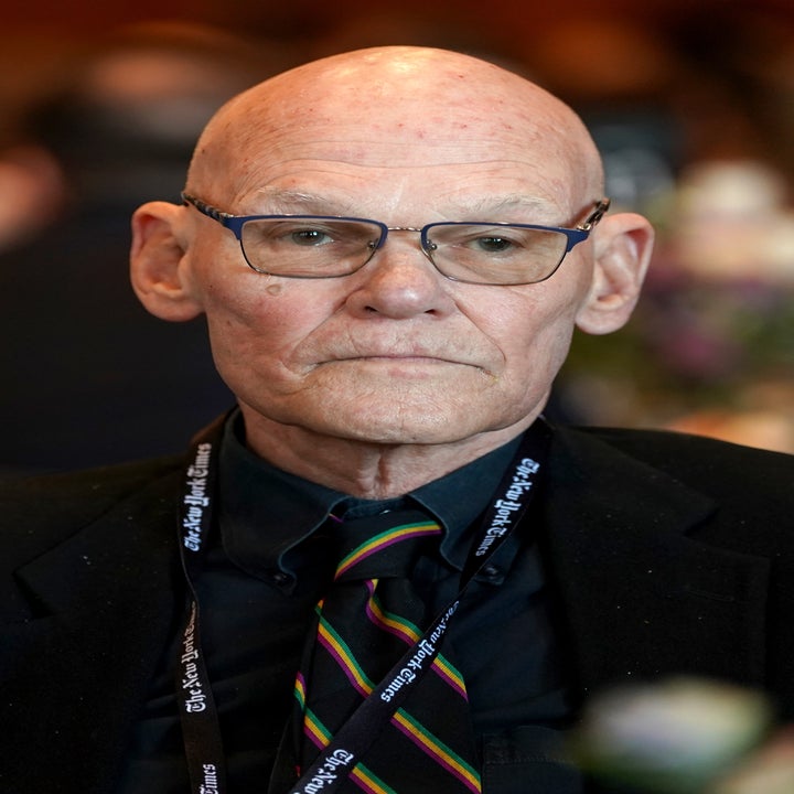 En person med briller og dress på en formell begivenhet, ser ettertenksom ut, omgitt av andre deltakere i bakgrunnen