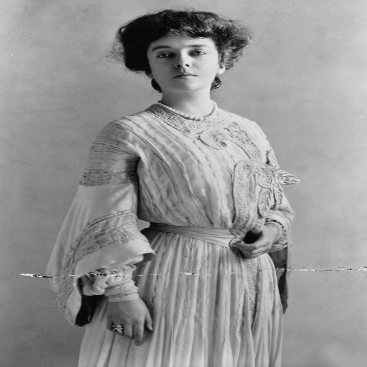 En kvinne fra tidlig på 1900-tallet iført en lang, elegant kjole med blondedetaljer, poserer formelt i studiomiljø