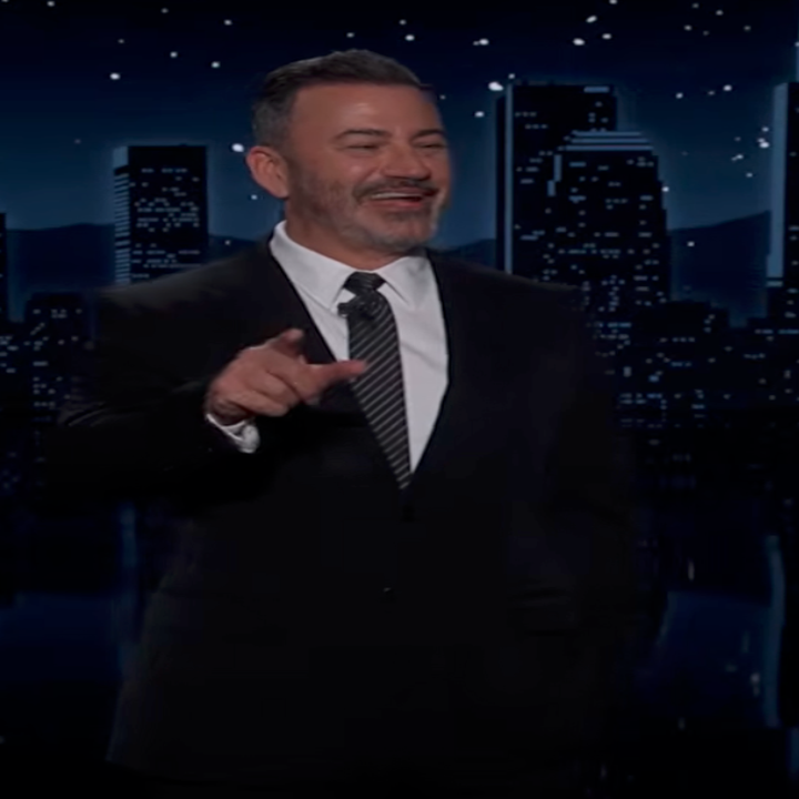 En person i dress snakker på et nattlig talkshowsett med en bysilhuett i bakgrunnen