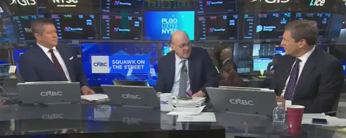 Tre nyhetsankere sitter ved en CNBC-pult og diskuterer finansielle nyheter i et børsrom. Monitorer viser ulike lagerdata
