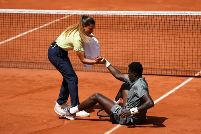 En tennisspiller sitter på banen og får hjelp til å reise seg fra en person i uformelt antrekk under en kamp