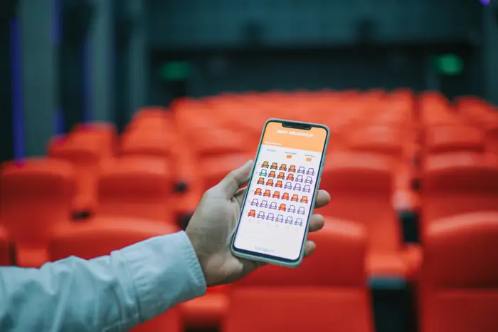 Person som holder en smarttelefon som viser en sittekartapp i et tomt teater, og illustrerer teknologiens rolle i arbeid og arrangementsplanlegging