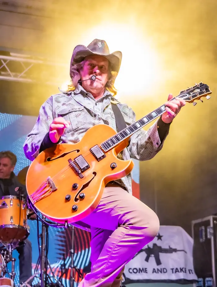 Musiker på scenen iført cowboyhatt og mønstret jakke, spiller energisk gitar under en liveopptreden