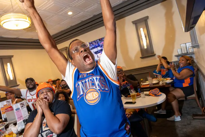 En gruppe entusiastiske basketballfans i en sportsbar, jubler med armene hevet. Man har på seg en Knicks-trøye, som legemliggjør spenning og lagånd