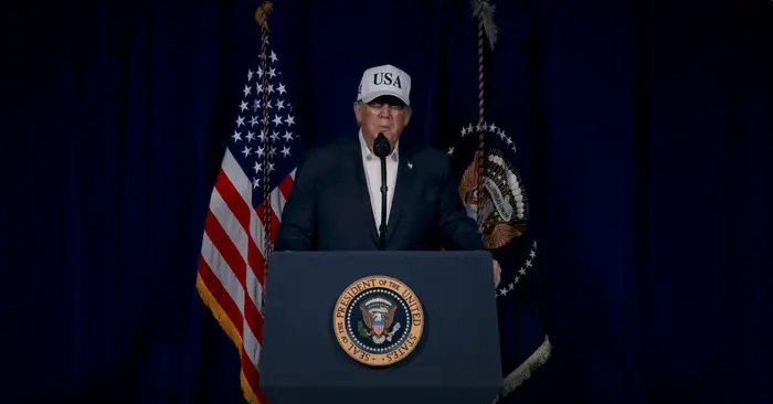 En person i dress og USA-hatt står ved et podium med presidentseglet, flankert av flagg