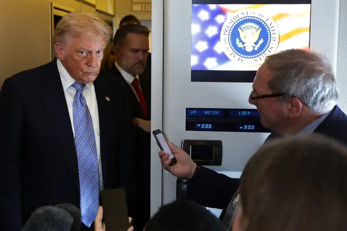 En person i dress og slips snakker med journalister på et fly. Presidentsegl og amerikansk flagg er synlig i bakgrunnen