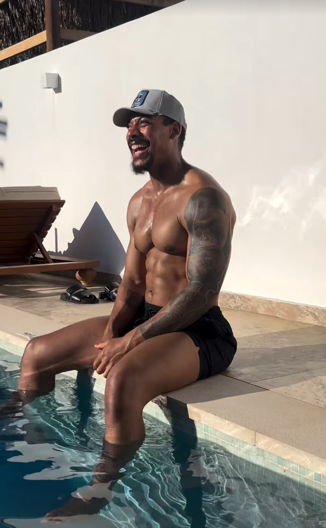Person som sitter ved et basseng, iført caps og shorts, smiler. De har en muskuløs bygning og slapper av på bassengkanten