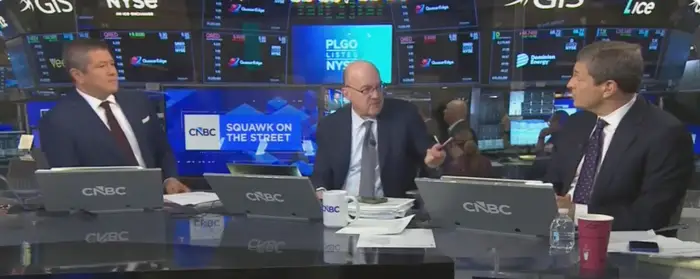 Tre CNBC-ankere sitter ved en nyhetsdisk og diskuterer børsoppdateringer; finansielle skjermer er synlige i bakgrunnen