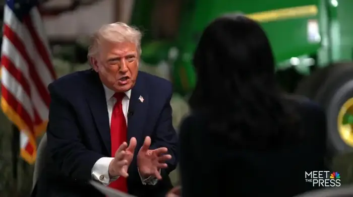 To personer er engasjert i et TV-intervju på "Meet the Press," med én som gestikulerer ettertrykkelig. Et amerikansk flagg og maskineri er synlige