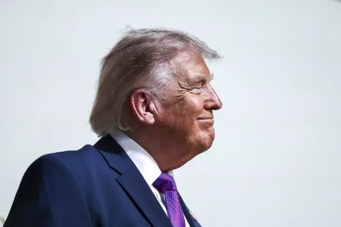 Donald Trump i dress og slips ser til siden, smiler lett mot en nøytral bakgrunn