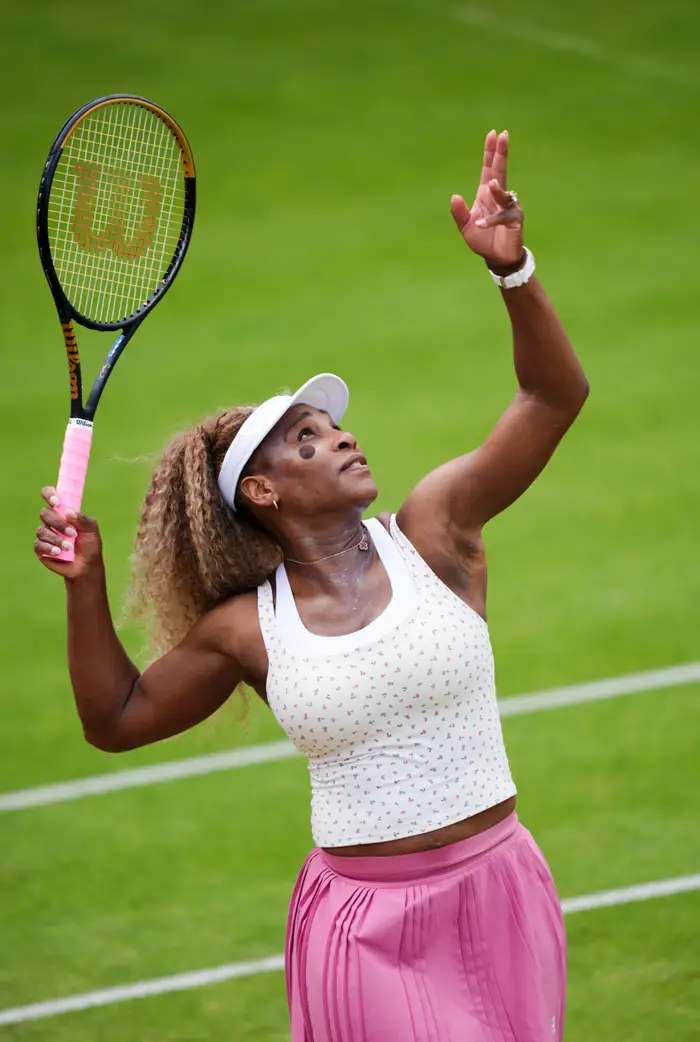En tennisspiller forbereder seg på å tjene på en gressbane, iført en hvit tank topp og rosa skjørt med visir