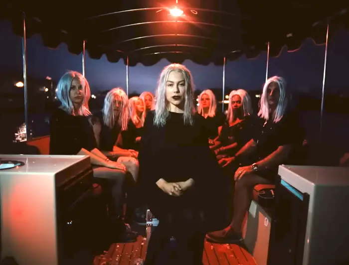 Folk med hvite parykker og mørkt antrekk sitter i en båt under svak belysning, og skaper en mystisk atmosfære
