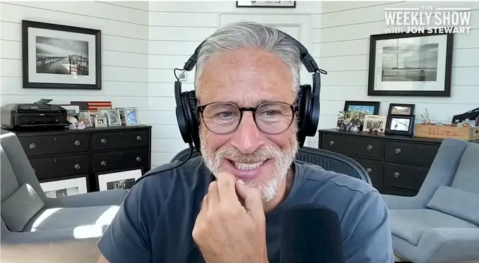 Person som har på seg hodetelefoner og briller, smiler mens han spiller inn en podcast på et hjemmekontor med innrammede bilder på veggen
