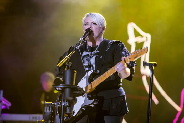 Person som opptrer på scenen med en elektrisk gitar, kledd i et svart antrekk. Mikrofon og musikkutstyr synlig