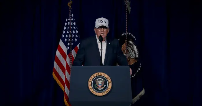 En person i dress og "USA" cap snakker på et podium med presidentseglet, flankert av flagg