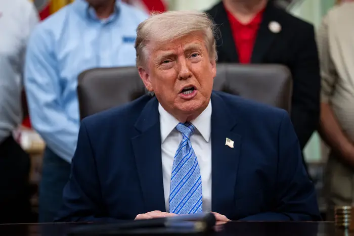 En person i dress sitter ved et skrivebord og snakker, med en amerikansk flaggnål på jakkeslaget. Andre står i bakgrunnen