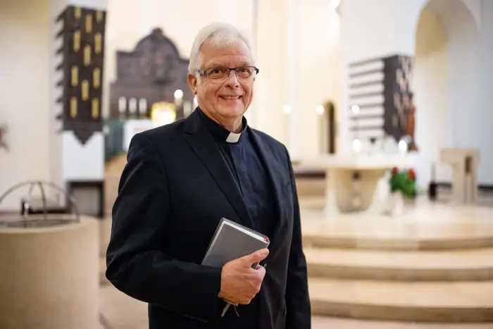 Et prestemedlem i svart dress holder en bok, stående i en kirke med moderne dekor