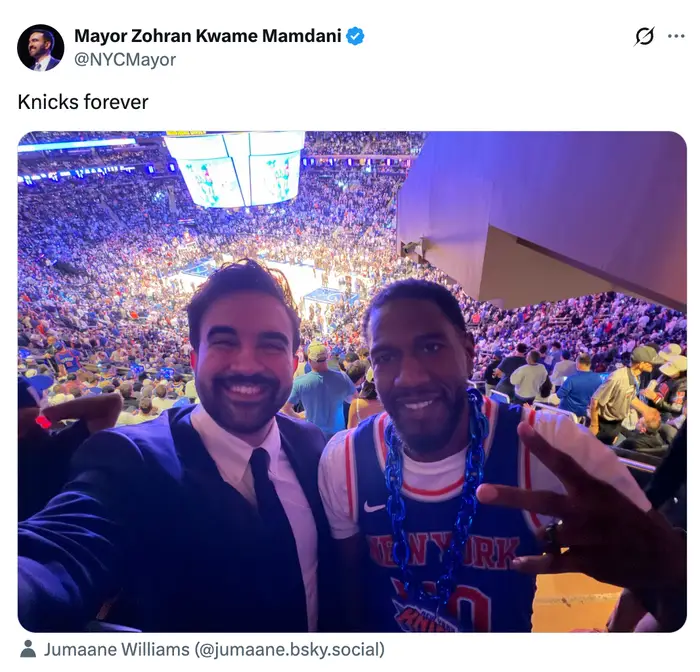 To personer som smiler, hvorav den ene har på seg en New York Knicks-trøye. De er på en fullsatt basketballkamp og tar en selfie fra tribunen