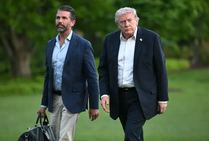 Donald Trump og Donald Trump Jr. går utendørs, den ene bærer en bag. Begge er i business casual antrekk. Frodige grøntområder er i bakgrunnen