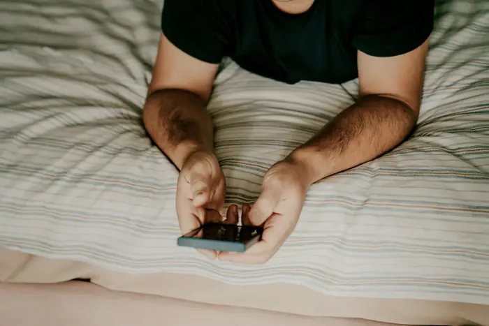 Person ligger på en seng og ser på en smarttelefon. Fokuset er på hendene og enhetene deres