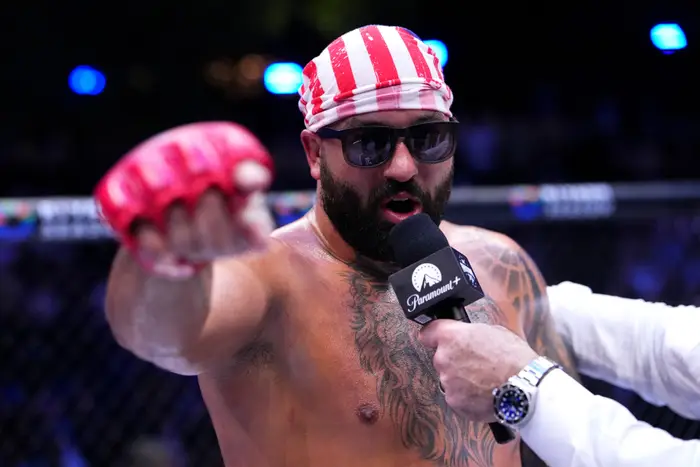 MMA-fighter i ringen, uten overkropp, iført solbriller og stripet bandana, snakker i en mikrofon mens han gestikulerer med en hanskebelagt hånd