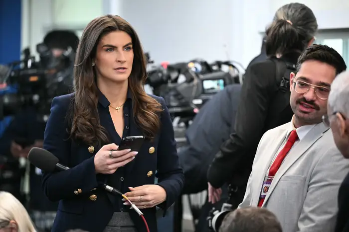 En person som holder en telefon står blant journalister og kameraer, iført en stilig jakke med gullknapper, ser ut til å være på en pressekonferanse