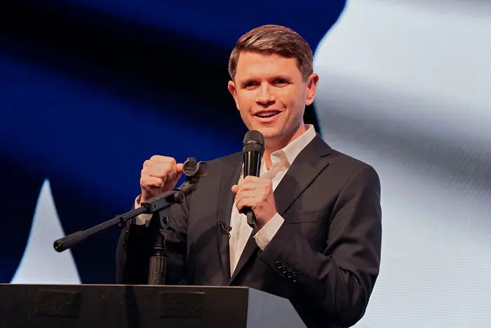 Person i dress snakker på et podium med en mikrofon, gestikulerer med en hånd foran et stort, uskarpt flaggbakgrunn