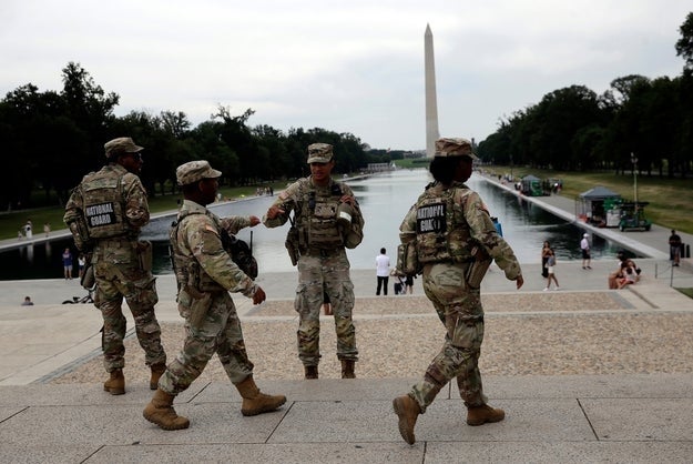 Fire uniformerte militærpersonell går nær det reflekterende bassenget med Washington-monumentet i bakgrunnen, blant besøkende og grøntområder