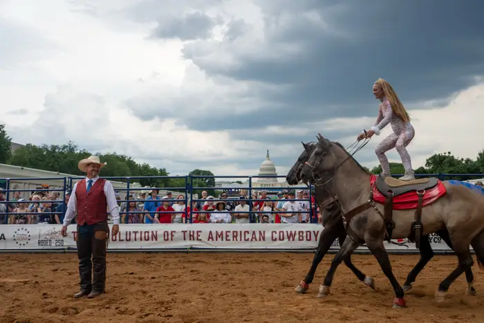 Person som står på hesteryggen og opptrer i en rodeo-arena med et publikum som ser på, cowboy observerer i nærheten
