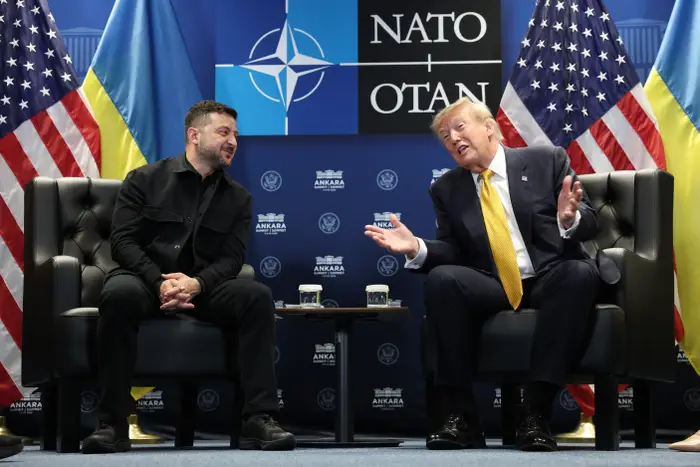To menn sitter på stoler og snakker energisk på et NATO-arrangement. Flagg er bak dem, og legger til kontekst til en diplomatisk møtesetting