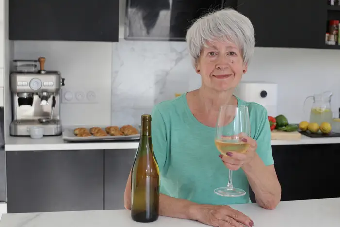 Person med kort hår på et kjøkken, som holder et glass hvitvin, smiler til kameraet; bak dem står bakevarer og en vinflaske
