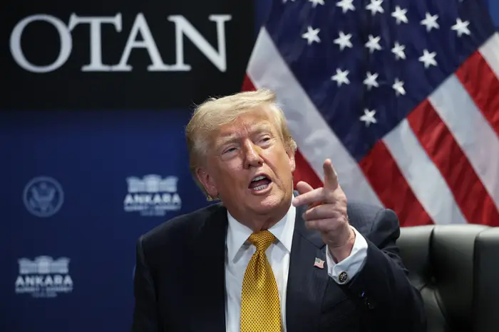 En person gestikulerer mens han snakker på et NATO-arrangement, og sitter foran et amerikansk flagg som bakgrunn