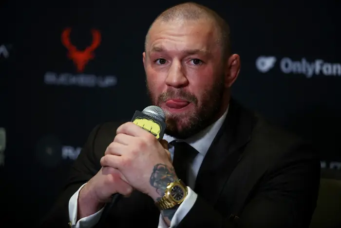 Conor McGregor i dress på en pressebegivenhet holder en mikrofon og virker gjennomtenkt