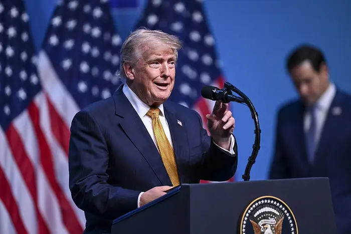 En person i dress snakker på et podium med amerikanske flagg bak seg, en annen person er uskarp i bakgrunnen