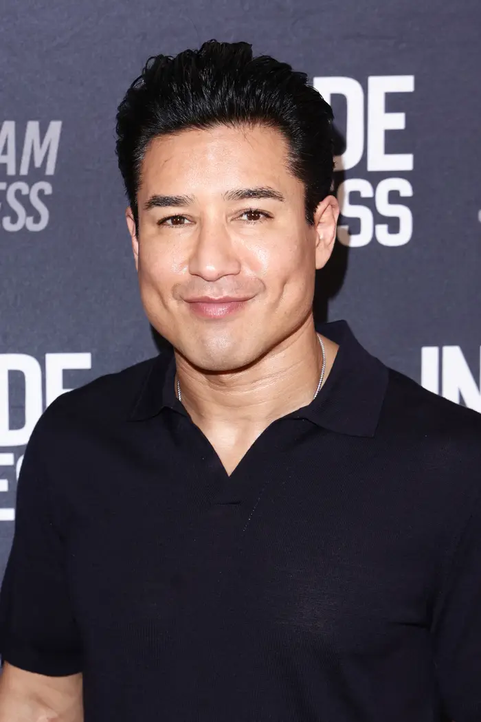 Mario Lopez smiler på et arrangement, iført en uformell skjorte, foran et bakteppe med tekst
