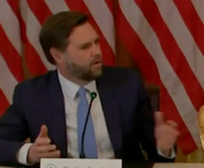En mann i dress gestikulerer mens han snakker på en konferanse. Amerikanske flagg er synlige i bakgrunnen