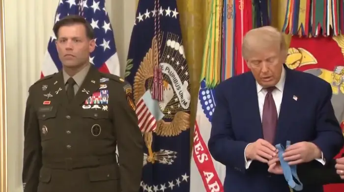 En mann i militæruniform står ved siden av en annen mann i dress og holder et medaljebånd ved en formell seremoni